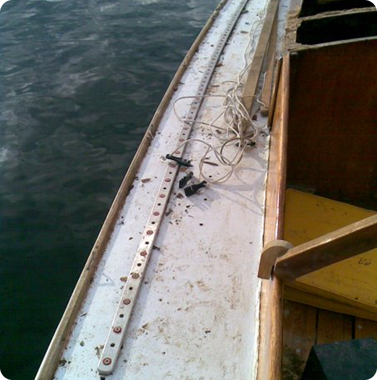 A close-up view of the inside edge of a small Skerry Cruiser, showing a white strip with circular markings, some rope, and wooden seats next to water.