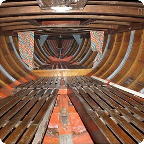 Interior view of a wooden Skerry Cruiser hull showing curved ribs, slatted floorboards, and colourful fabric lining parts of the walls. The image is taken from the centre, facing towards the bow.