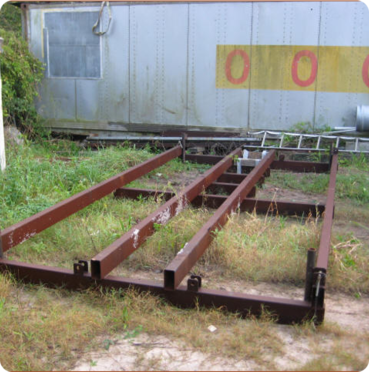 A steel frame structure with four parallel beams sits on overgrown grass beside a weathered metal building marked with yellow and red circles—a scene reminiscent of the careful restoration of Classic Sailing Yachts. A ladder leans against the building in the background.