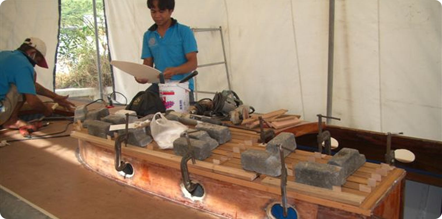Two people work on a wooden Square Metre Yachts model under a marquee. Tools, clamps, and bricks are arranged on the boat, with one person holding a white object as the other works in the background.