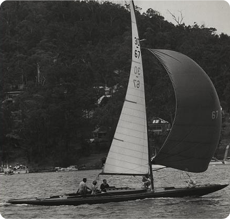 A black and white photo of three people sailing a sleek Schärenkreuzer, or Skerry Cruiser, with the number 67 on its sail, gliding across the water with a wooded shoreline and houses in the background.
