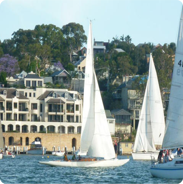 Square Metre Yachts with tall white sails glide on calm water in front of large waterfront houses and green, tree-covered hills on a sunny day.