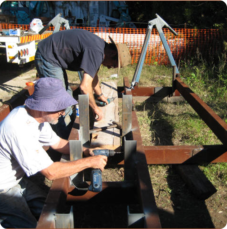 Two people wearing hats use power drills to work on a metal frame structure outdoors, possibly assembling parts for Classic Sailing Yachts, with orange safety fencing and construction materials in the background on a sunny day.