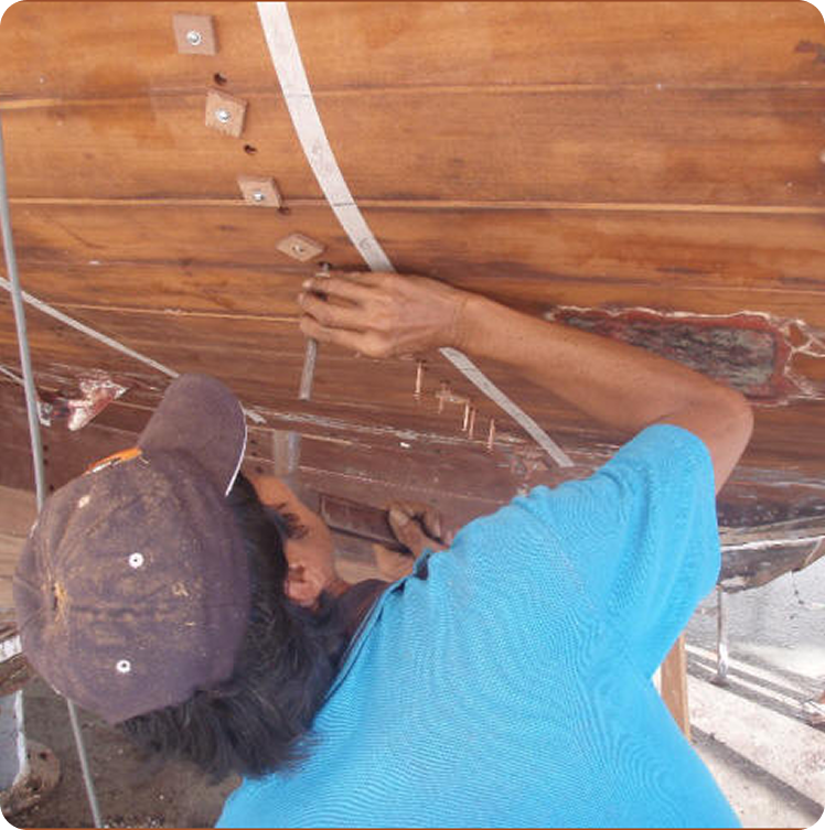 A person in a blue shirt and cap repairs the wooden hull of a Classic Sailing Yacht, attaching metal strips and fasteners to the underside, working carefully with both hands.