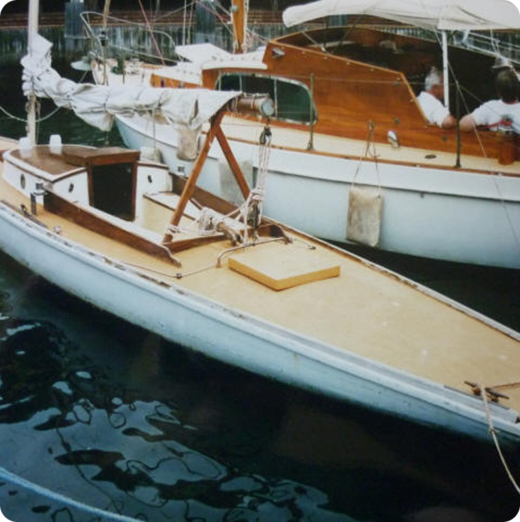 A small Skerry Cruiser sailboat with a tan deck and white hull is moored on calm water next to a larger wooden cabin boat. The sailboat’s mast is lowered, with ropes and equipment scattered on its deck.