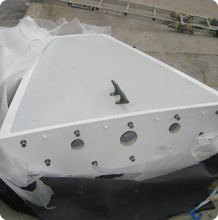 A large, white, triangular metal panel with circular holes and bolts, likely from a Square Metre Yachts or Schärenkreuzer project, lies flat on the ground. A metal cleat is attached near the centre, with plastic sheeting partially covering the sides.