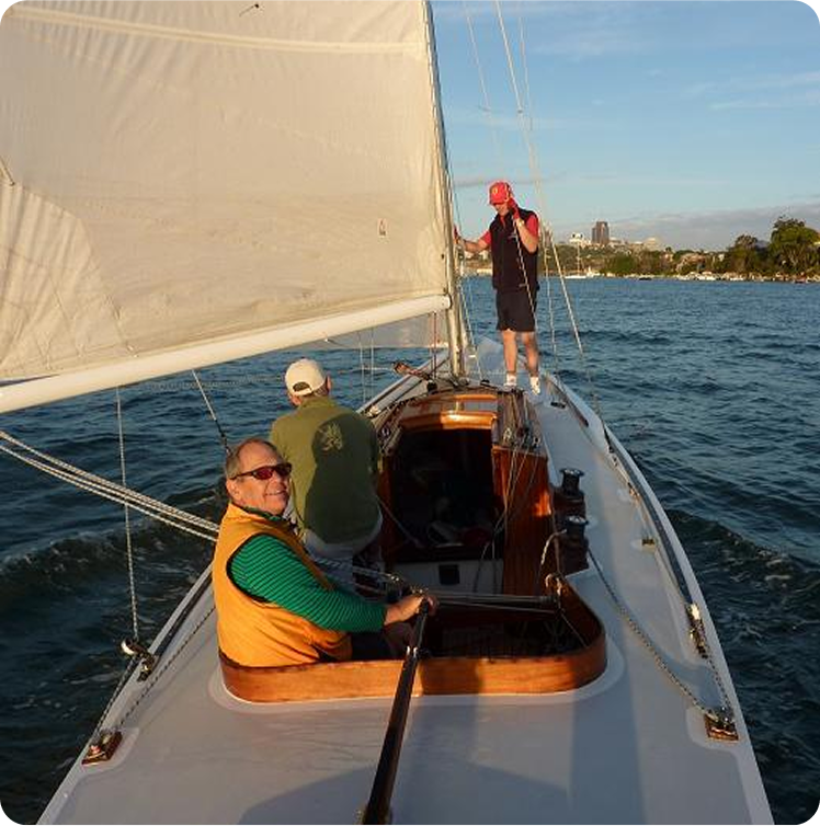 Three people are sailing on a white Skerry Cruiser. One in a yellow vest is steering, another sits facing forwards, and the third stands at the bow waving. The water is calm and land is visible in the background.