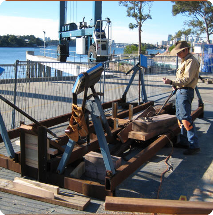 A person in outdoor work gear examines a timber structure with blue metal supports on a dock, near classic sailing yachts and Schärenkreuzer moored by the water, with trees and buildings in the background.