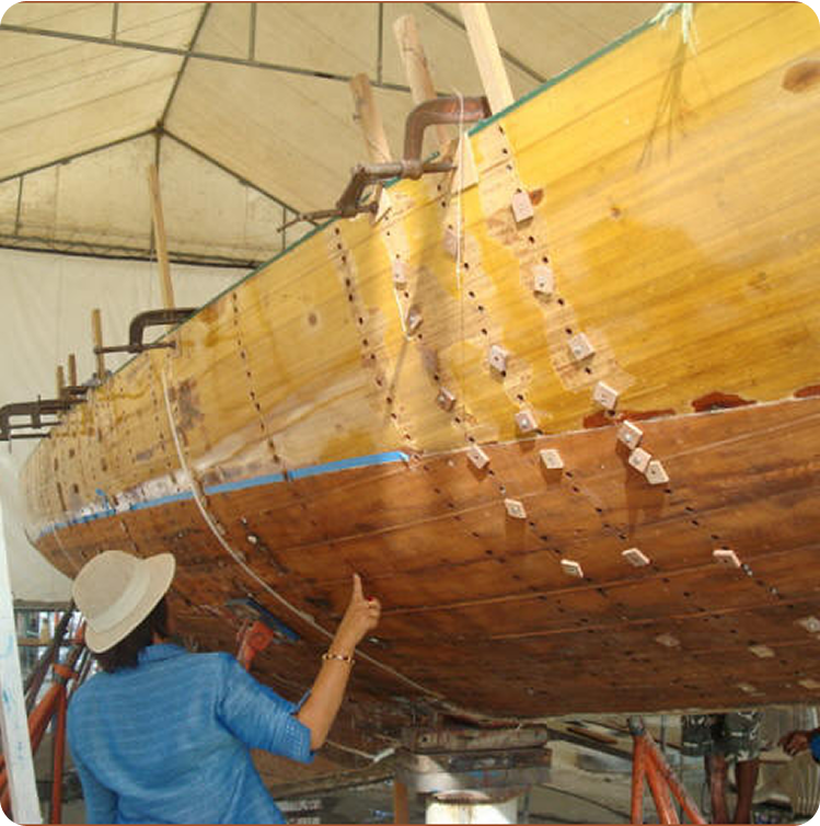 A person wearing a sunhat examines the wooden hull of a Classic Sailing Yacht under construction or restoration inside a marquee, with clamps and tools attached to the boat’s surface.