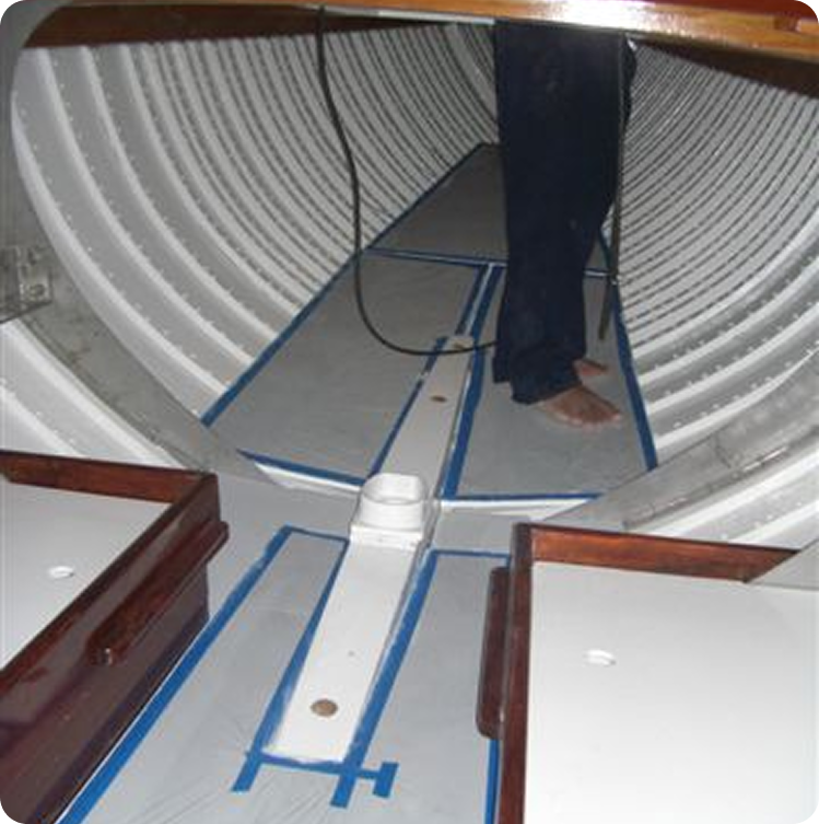 Person standing barefoot inside a boat hull with curved ribbed walls, wooden and white panels on the floor, and blue tape marking sections—a scene reminiscent of Classic Sailing Yachts. A cable hangs from above.