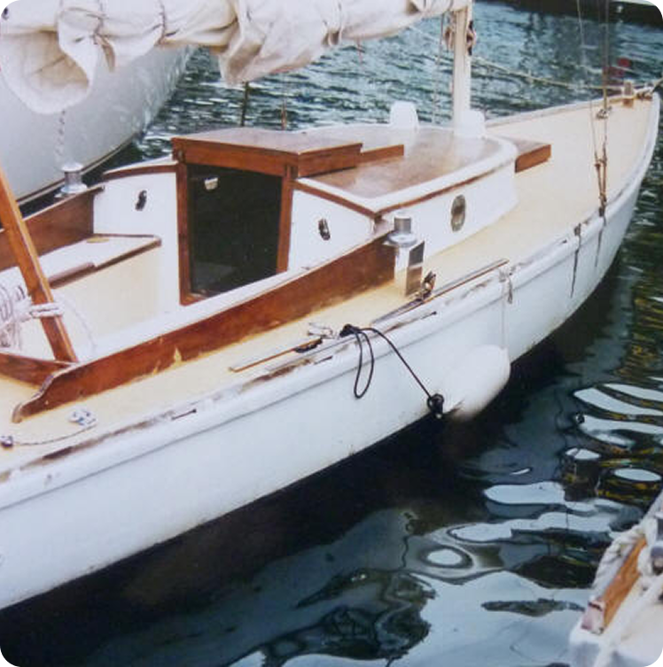 A small wooden sailing boat, reminiscent of Classic Sailing Yachts, with white sides and a tan deck floats on calm water. The boat has a cabin with wooden trim, a furled sail on the boom, and a white fender hanging over the side.