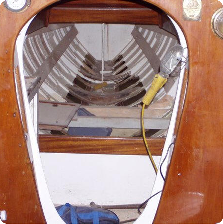 View through a wooden boat’s oval hatch showing the empty, ribbed interior hull—a scene reminiscent of classic Skerry Cruisers. A hanging work light with a yellow handle and an electrical flex is visible on the right side of the hatch frame.