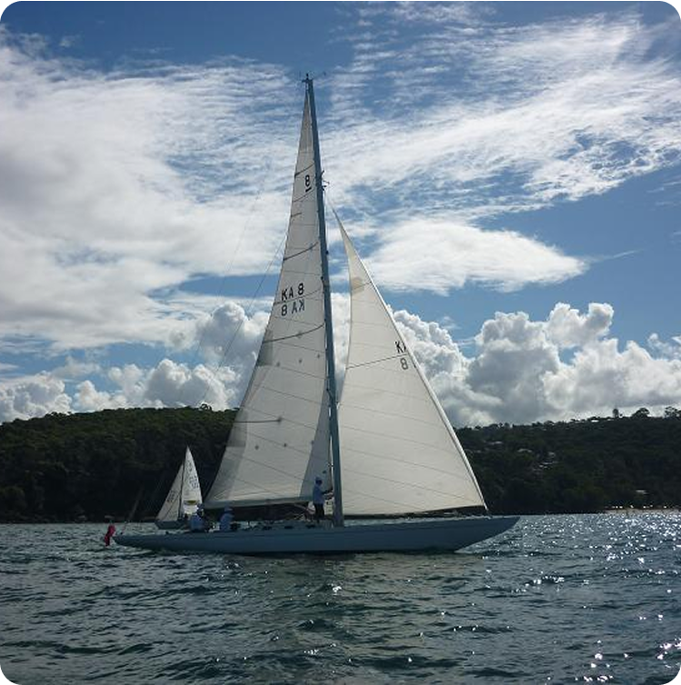 A classic sailing yacht with white sails glides on the water under a partly cloudy sky, with green, tree-covered hills in the background.