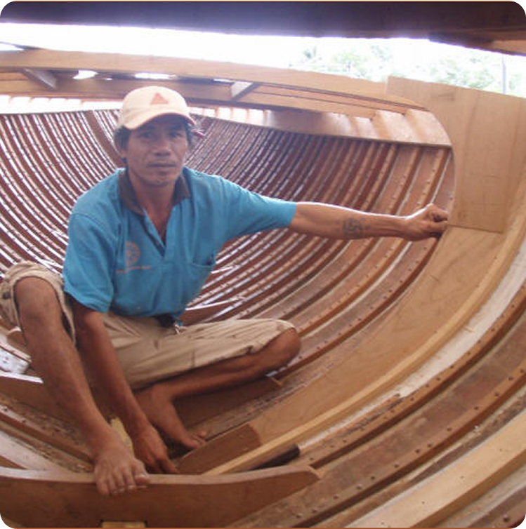A man in a blue shirt and cap sits barefoot inside a classic sailing yacht under construction, holding a wooden piece against the boat’s frames as sunlight filters through the open top.