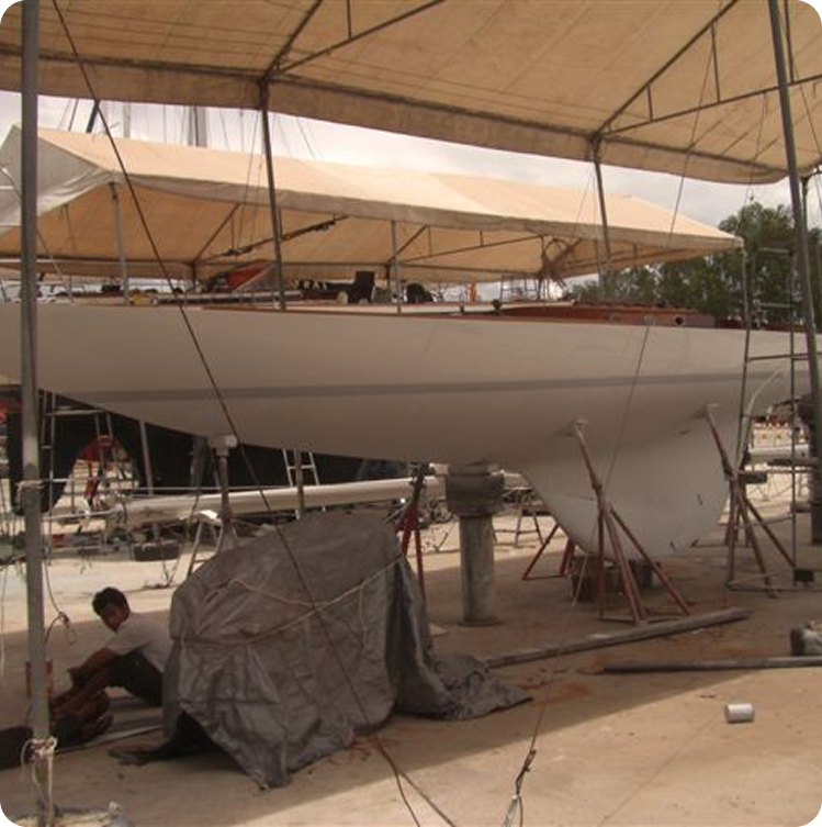 A white Square Metre Yacht is being worked on in a covered outdoor area. Tarpaulins and scaffolding surround the Skerry Cruiser, whilst a person crouches nearby, appearing to perform maintenance or repairs.