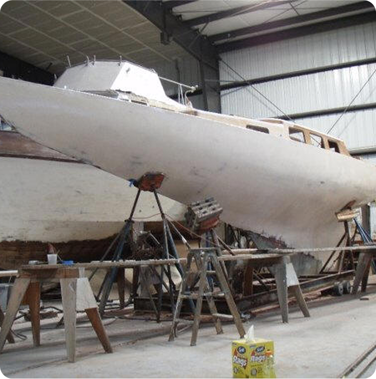 A Schärenkreuzer sailboat is propped up on supports inside a boatyard or workshop. The classic sailing yacht’s hull appears to be under restoration, with tools and workbenches nearby in the indoor space.