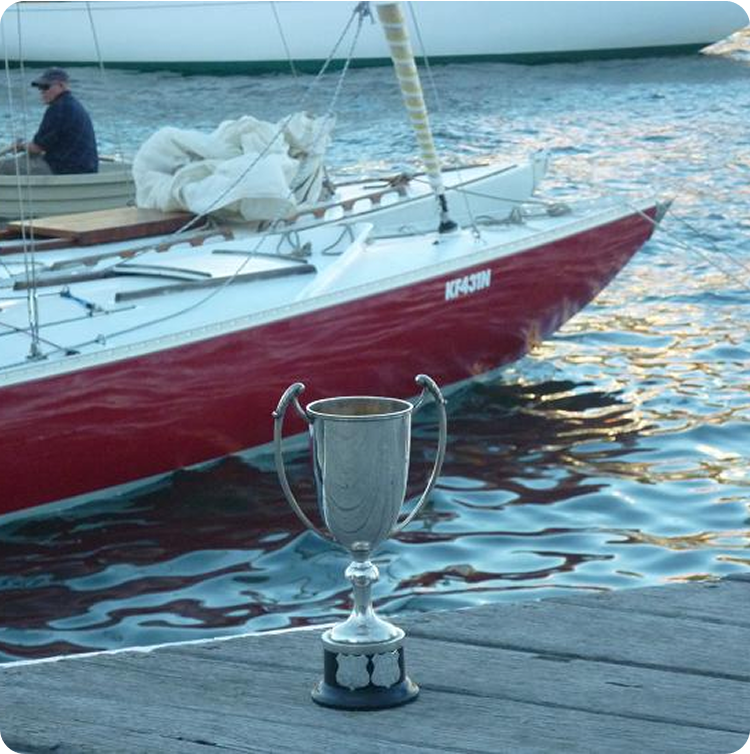 A silver trophy cup sits on a wooden jetty in front of a classic Square Metre Yacht moored in the water, with a person seated on the boat.