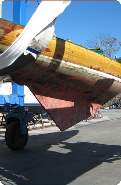 A close-up view of the underside of a Classic Sailing Yacht on land, showing a red, weathered fin keel and part of a wheel from the boat stand, with blue sky and a few trees in the background.