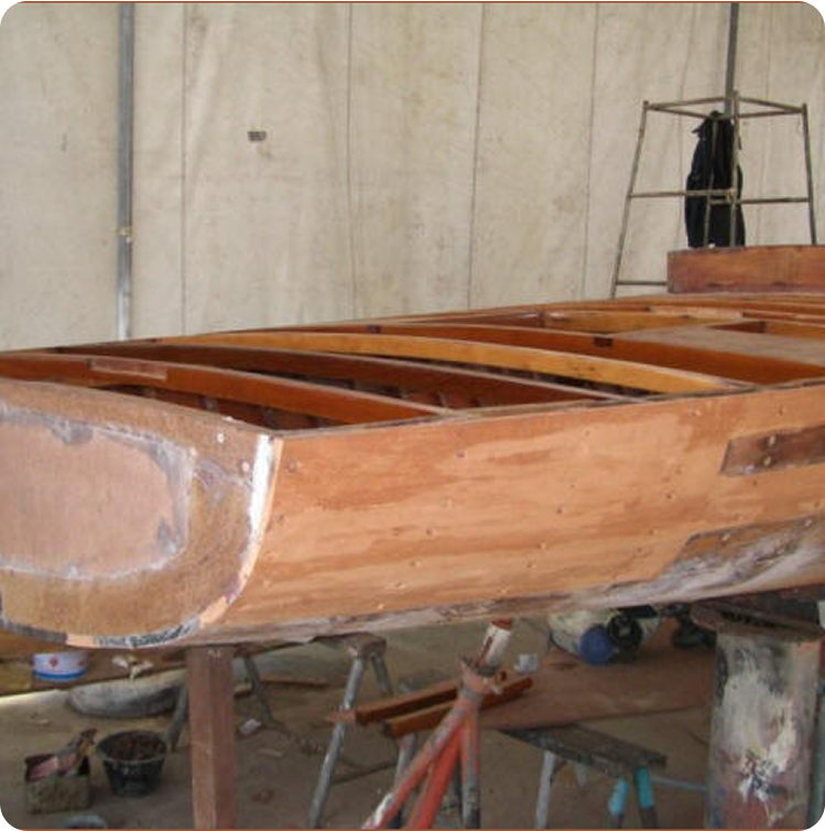 A wooden Skerry Cruiser under construction in a workshop, showing the unfinished hull and exposed internal framework, with tools and equipment visible in the background.