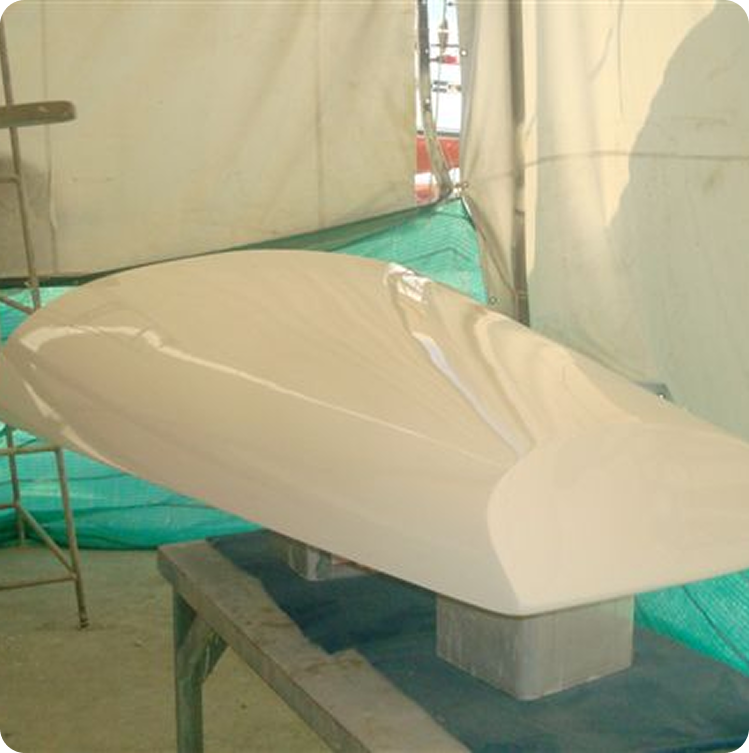 A smooth, cream-coloured boat hull is positioned upside down on a workbench inside a workshop, reflecting the classic lines of Square Metre Yachts. Green mesh and white fabric panels add contrast in the background.