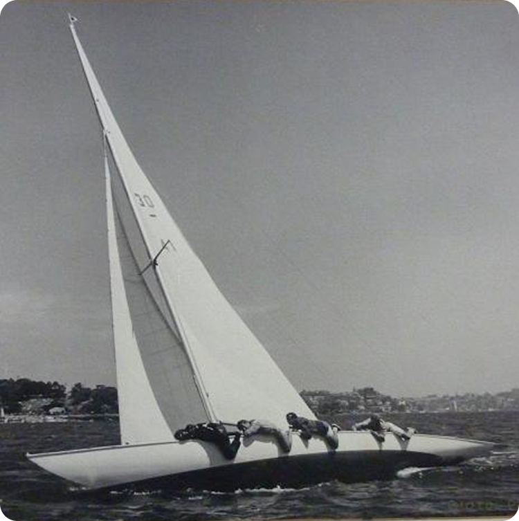 Black and white photo of a classic Square Metre Yacht leaning sharply on the water, with several crew members hanging off the side for balance. The shoreline and trees are visible in the background.