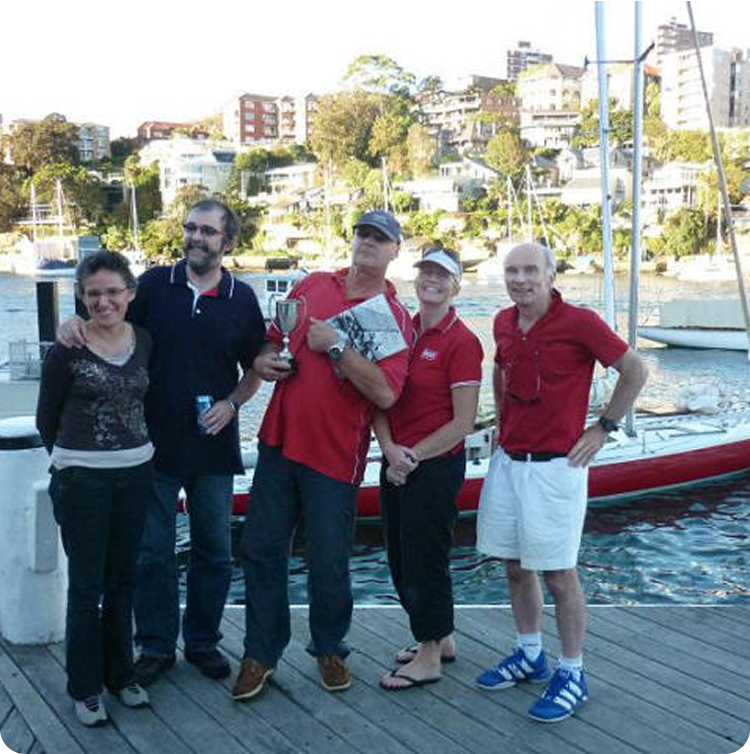 Five people stand on a wooden jetty by the water, smiling at the camera. One person holds a trophy. Classic sailing yachts and waterfront houses are visible in the background. It appears to be a sunny day.