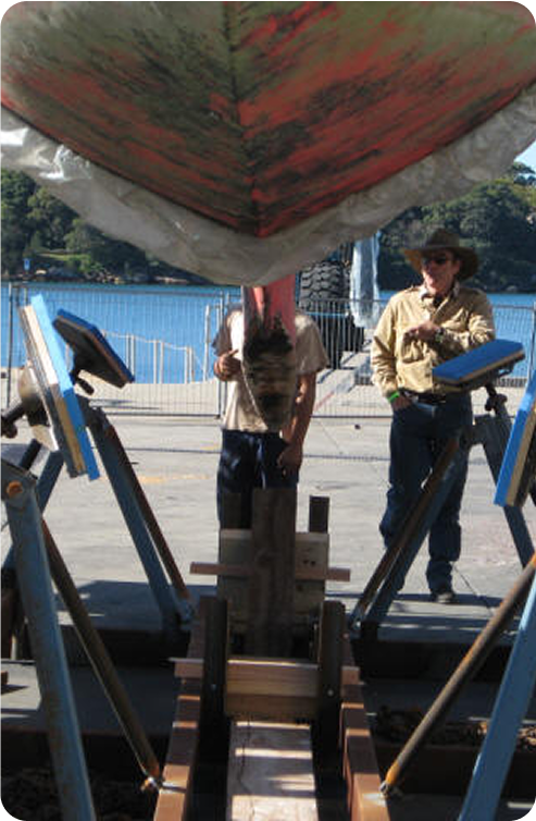 Two people stand beneath the hull of a Classic Sailing Yacht propped up on supports outdoors near water, with trees visible in the background. One person is partially obscured by the boat’s keel.