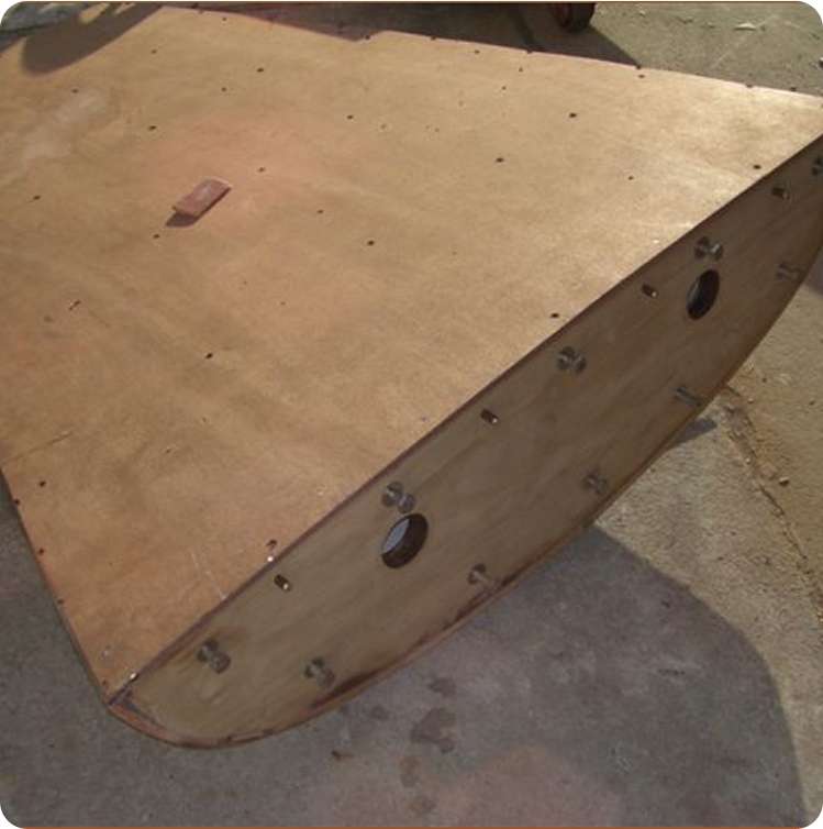 A close-up of a wooden Schärenkreuzer hull under construction, showing a flat surface with rounded edges, circular cut-outs, and multiple fasteners, resting on a concrete floor.