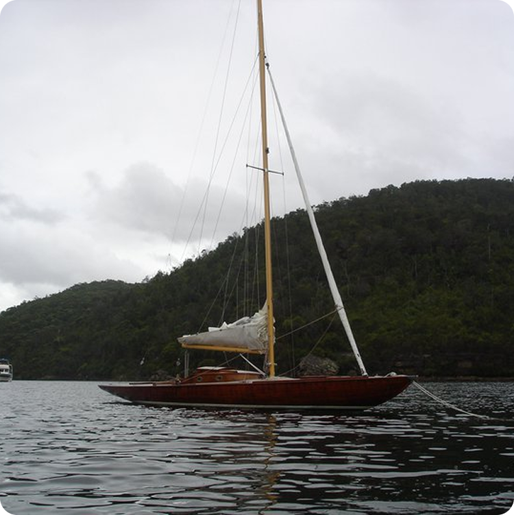 A classic wooden sailing boat, reminiscent of Skerry Cruisers, floats on calm water with its sail down, anchored near a tree-covered hillside under a cloudy sky.