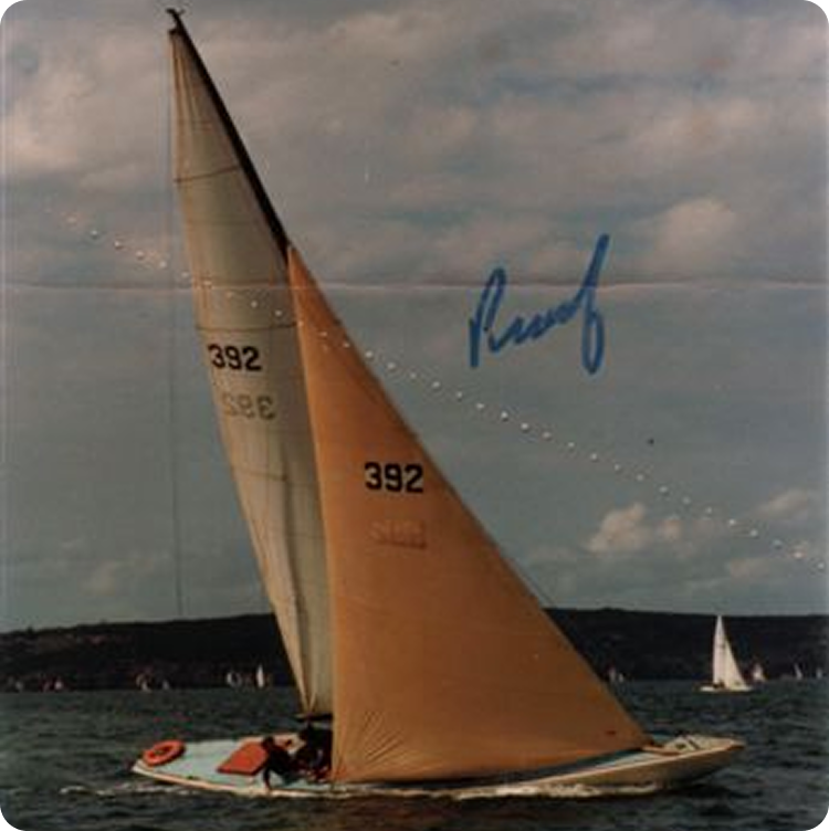 A sailboat with tan sails marked 392, likely a classic Schärenkreuzer or Square Metre Yacht, glides on the water under a partly cloudy sky; distant land and other sailboats are in the background. A blue signature is written across the sky.