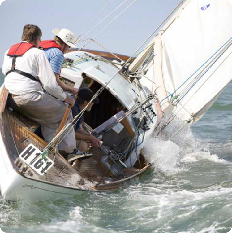 Three people in life jackets sail a small wooden Skerry Cruiser, leaning as the boat tilts sharply and cuts through choppy water, creating splashes near the prow.