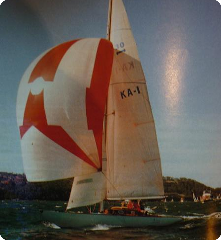 A sailing boat with the number KA-1 on its sail glides across the water with its colourful red and white spinnaker deployed. Among classic sailing yachts, it stands out as other boats and trees are visible in the background under a blue sky.