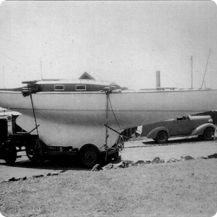 A black-and-white photo showing a classic Schärenkreuzer sailboat secured on a trailer, parked near a vintage car. The background reveals rocks, grass, and the faint outlines of buildings and boats.