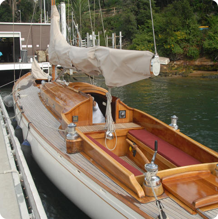 A classic wooden Schärenkreuzer sailboat with a covered mast is moored at the dock. The boat features polished wood details, red cushioned seating, and sits beside green water with trees and a building in the background.