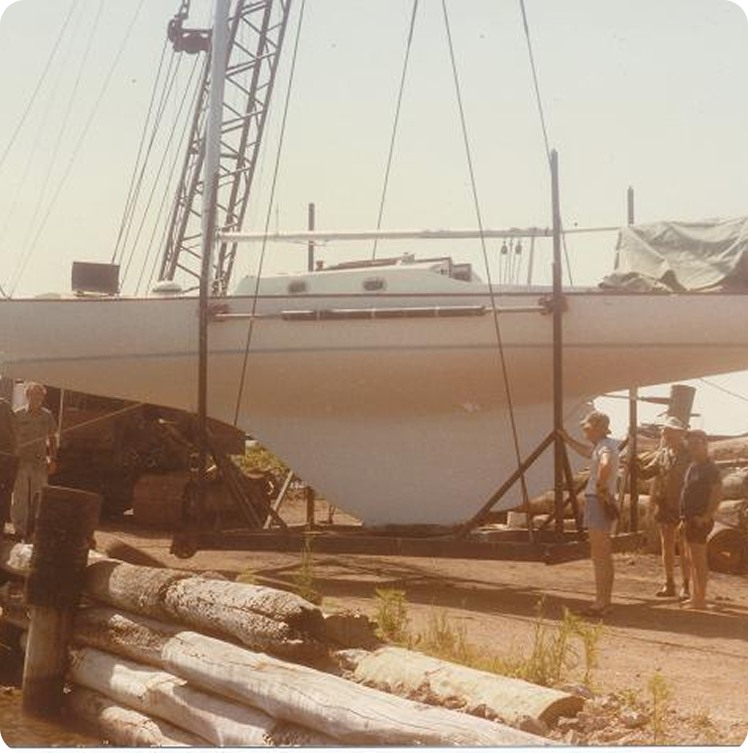 A sailboat, likely a classic Schärenkreuzer, is suspended by a crane above the ground near a quay, with several people standing nearby and logs piled in the foreground. The sky is clear and sunny.