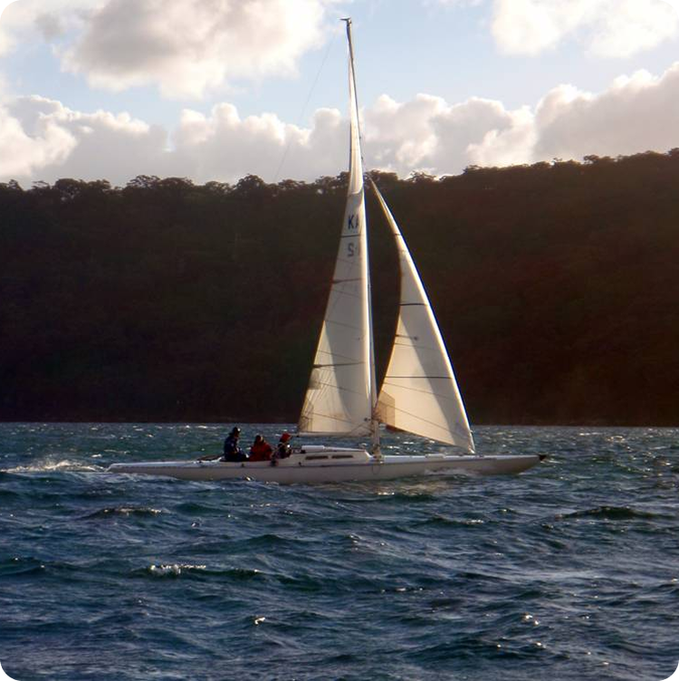 A classic sailing yacht with several people on board glides across choppy blue water, with forested hills and a cloudy sky in the background.