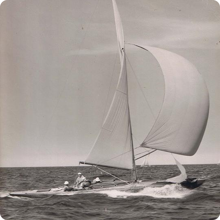 A black-and-white photo of a Classic Sailing Yacht with a tall mast and billowing sails gliding quickly across the open sea, with several people on board. The sky is mostly clear.