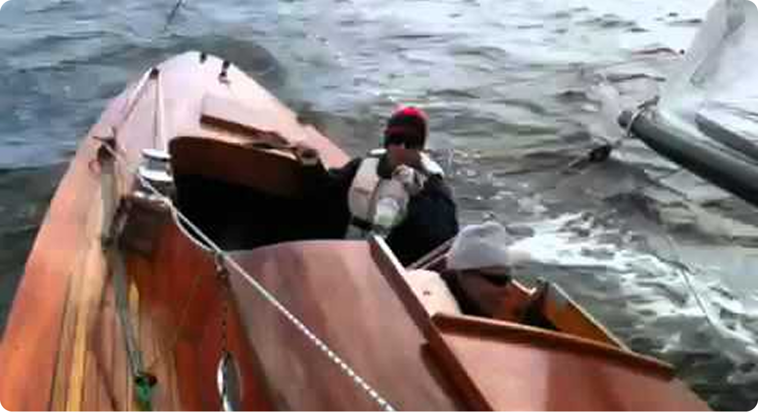 Two people in life jackets sit in a small wooden sailing boat reminiscent of Classic Sailing Yachts on a wavy body of water, with one person partially inside the cabin and another holding onto the edge.