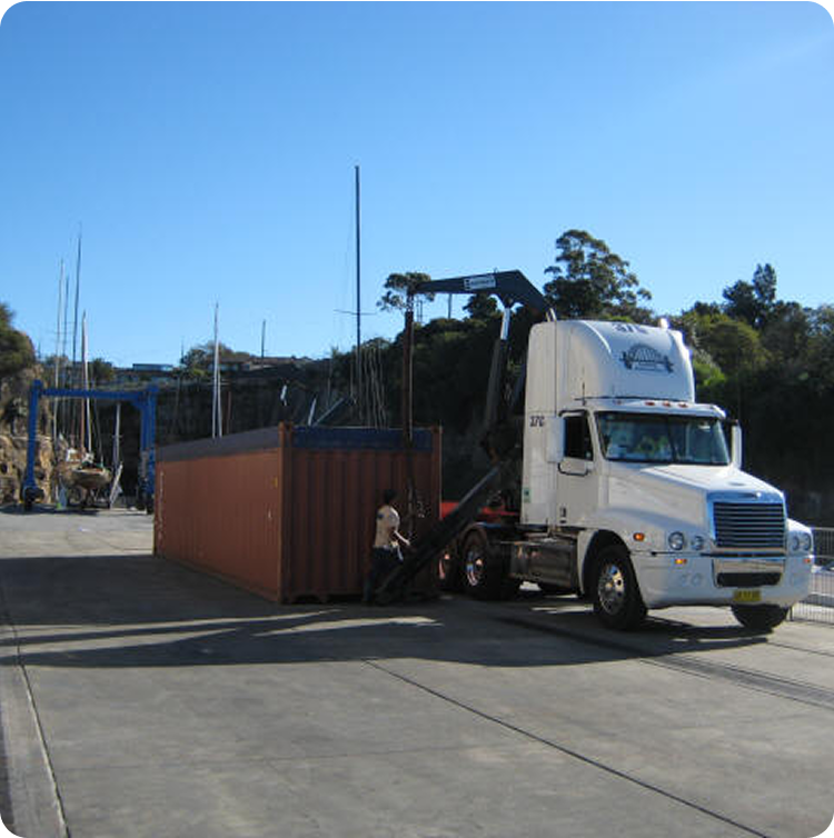 A white lorry is loading or unloading a large brown shipping container with a crane at an industrial dockyard, with Skerry Cruisers and classic sailing yachts visible near boats and trees under a clear blue sky.