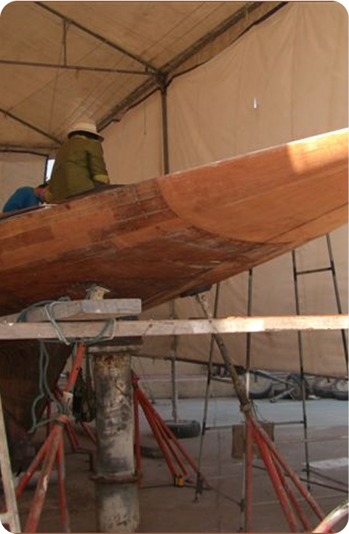 A person works on the wooden hull of a Classic Sailing Yacht inside a tented workshop, with ladders, scaffolding, and tools visible around the vessel.