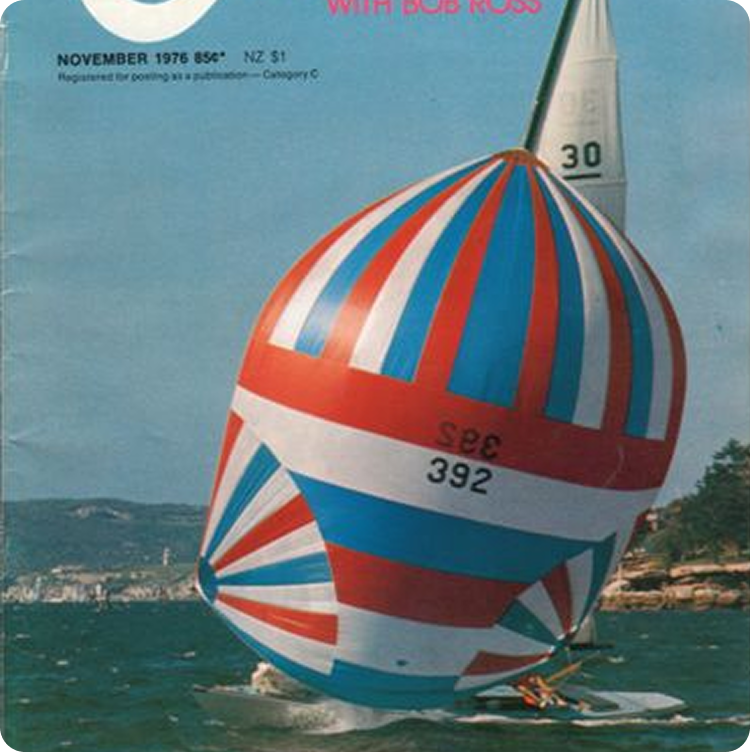 A classic sailing yacht with a large, colourful red, white, and blue spinnaker sails close to shore on the cover of a November 1976 magazine. Water, hills, and trees are visible in the background.