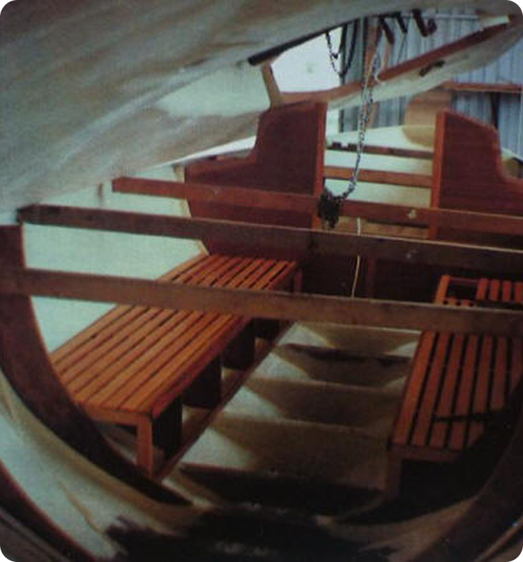 Interior view of a small wooden boat, typical of classic Square Metre Yachts, showing two wooden benches on either side, steps leading up, and panelling with light streaming in from the open hatch above.
