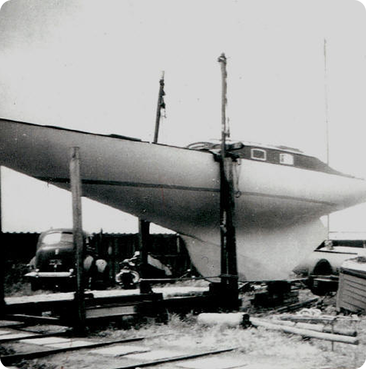 Black and white photo of a Skerry Cruiser sailboat elevated on supports in a yard, with vintage cars and assorted equipment nearby. The boat, a classic sailing yacht, is viewed from the side, showing its hull and cabin.