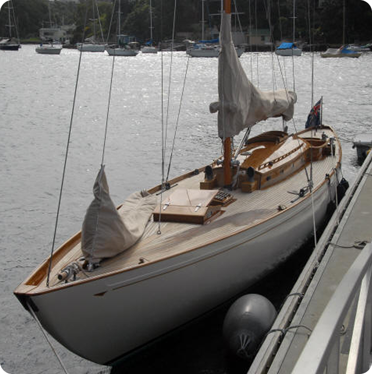 A wooden Skerry Cruiser with covered sails is moored at a pier on a calm body of water, with other boats and trees visible in the background.
