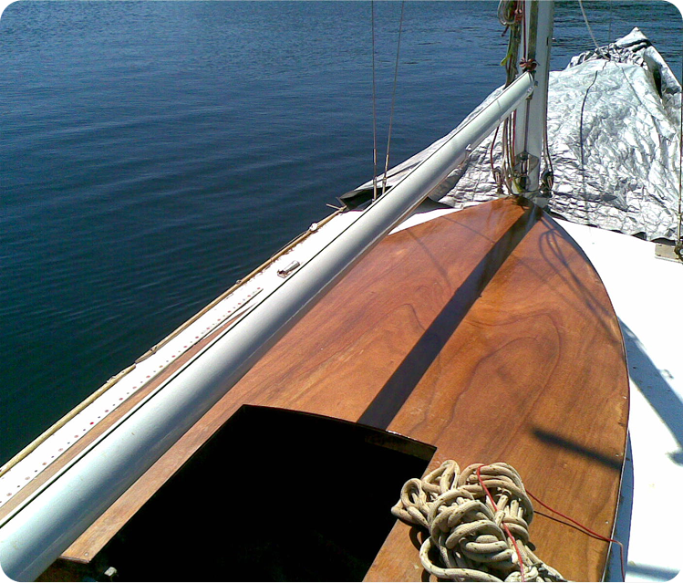 A close-up view of a Schärenkreuzer sailboat deck with polished wood, a coiled rope, and part of the mast visible, floating on calm blue water under bright sunlight.