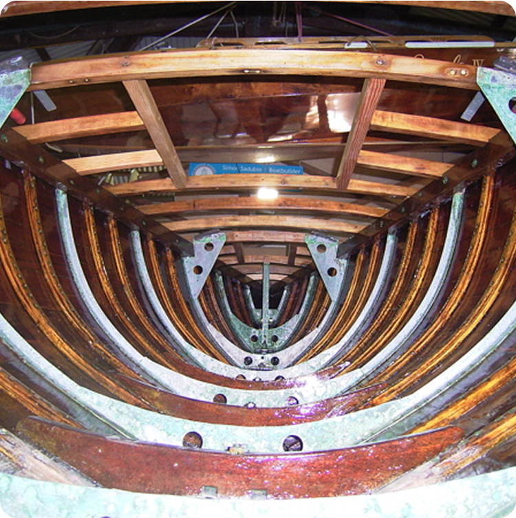 The image shows the interior framework of a wooden boat under construction, highlighting curved ribs, metal brackets, and wooden crossbeams viewed from the stern towards the bow—typical of classic sailing yachts or Schärenkreuzer craftsmanship.