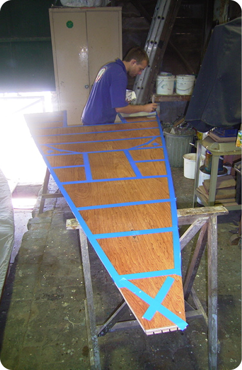 A person works on a wooden boat deck resting on sawhorses, with blue painter’s tape marking sections—evoking the elegance of Classic Sailing Yachts. Tools, paint tins, and a ladder are visible in the busy workshop background.