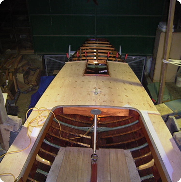 A wooden Skerry Cruiser is under construction in a workshop, with planks partially installed on the deck and open ribbing visible inside the hull. Tools and materials are scattered around the area.