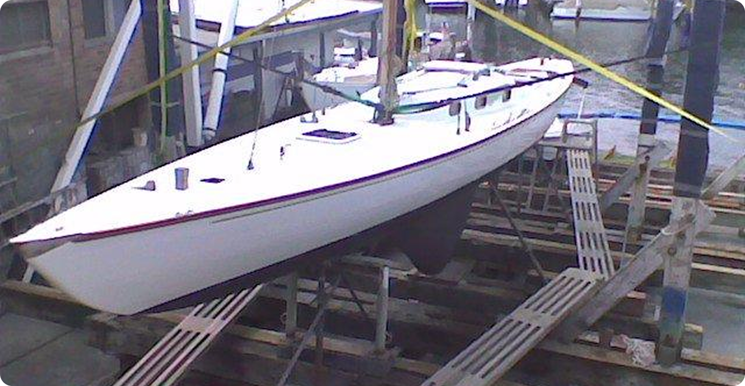 A classic sailing yacht, possibly a Skerry Cruiser, is positioned on wooden supports at a dockyard, elevated above the water for maintenance or repairs. Yellow straps secure the boat, and a building is visible alongside the quay.