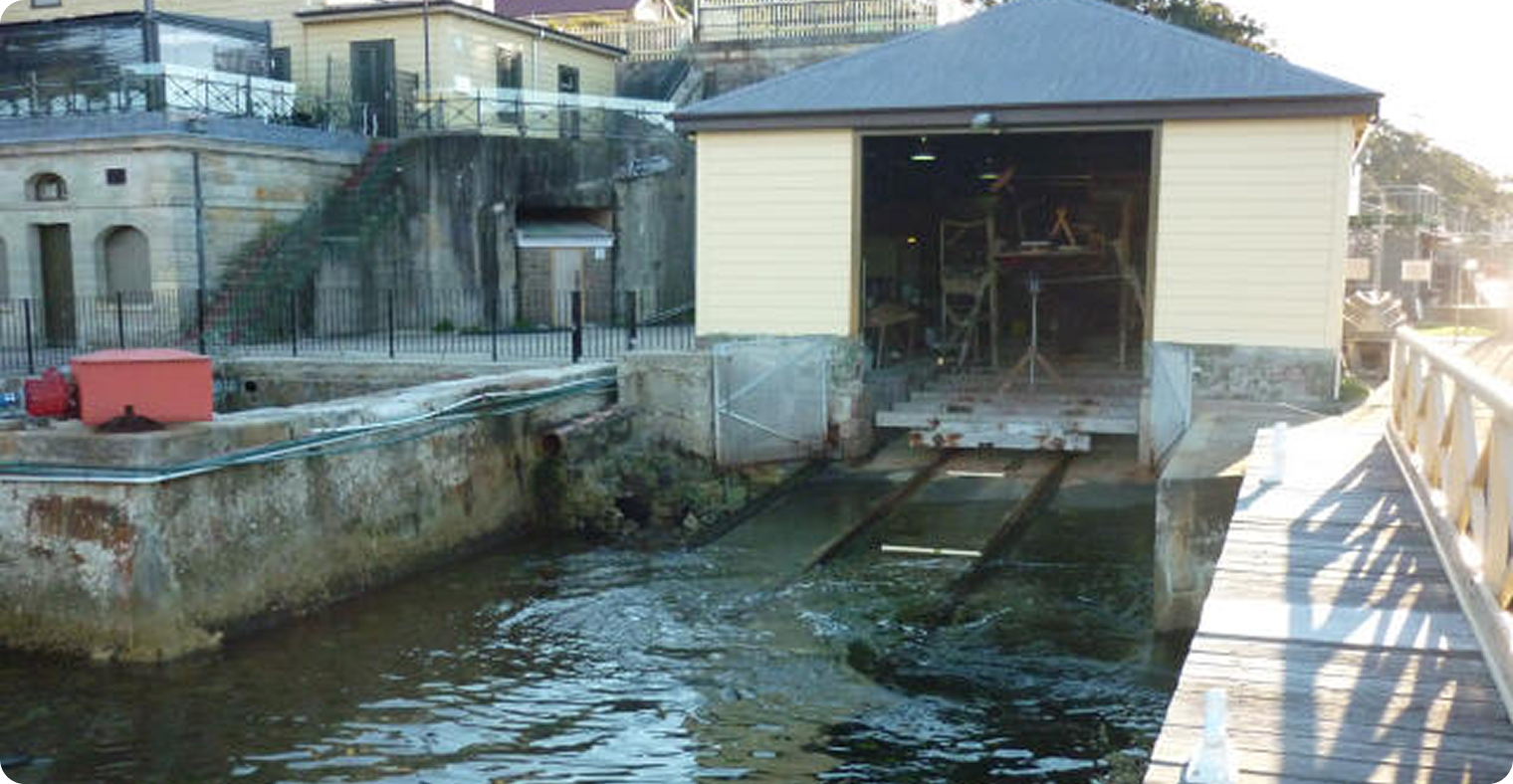 An old boat shed with an open entrance, situated beside the water, once a haven for Skerry Cruisers. Rails extend from the shed into the water, with stone walls, a walkway, and historic buildings in the background.